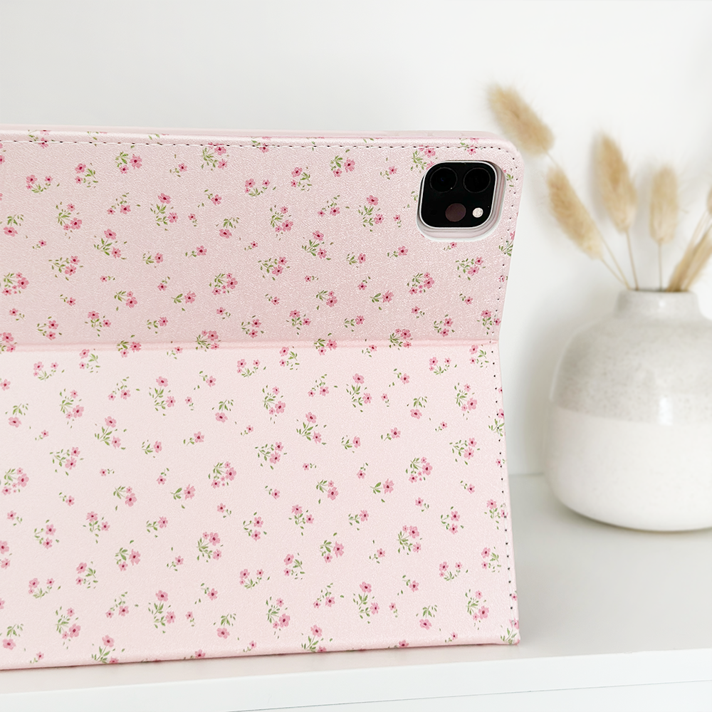 The Ditsy Floral Pink iPad Case from Coconut Lane, adorned with small floral patterns, is displayed open on a white surface. In the background, a ceramic vase holds a few dried pampas grass stems, adding a touch of natural decor. Crafted from vegan leather, this Apple iPad case combines elegance with sustainability.