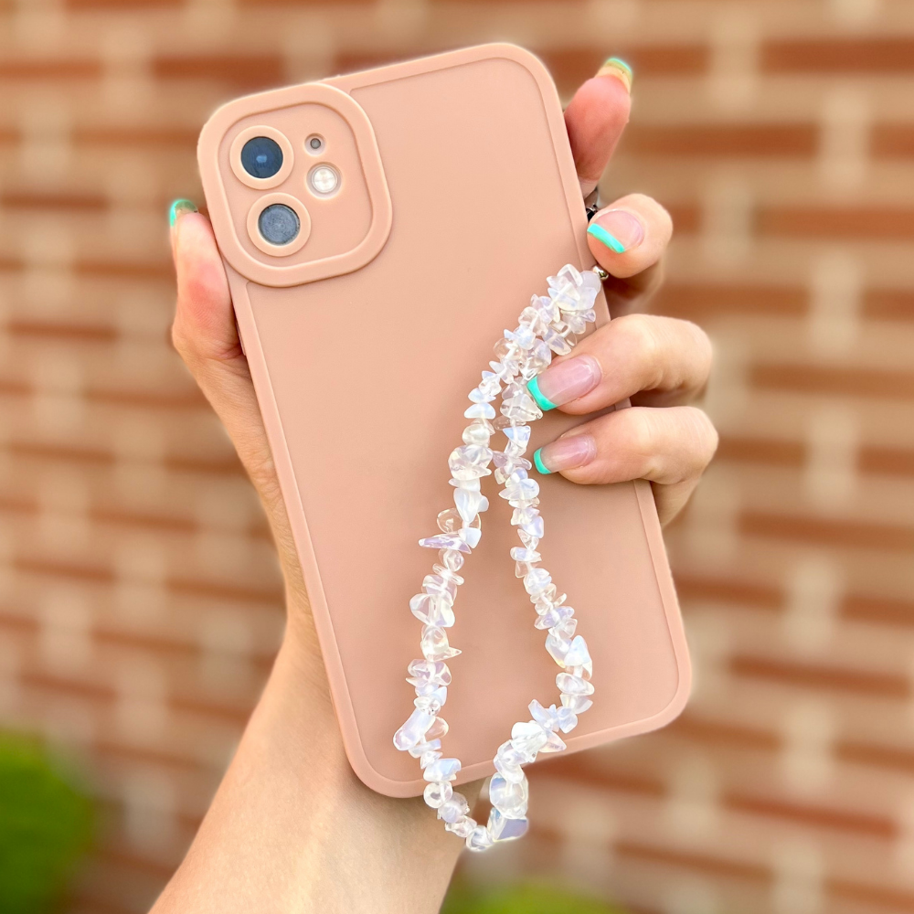 Pink phone with Coconut Lane Crystal Moonstone phone strap held by hand with colorful nails; blurred brick wall background.