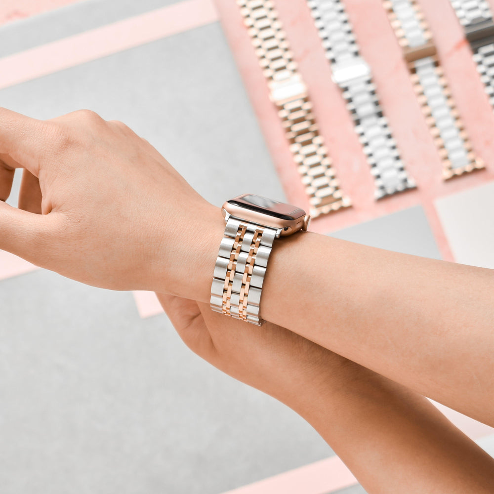 A wrist model displays the Coconut Lane Stainless Steel Apple Watch Strap - Silver & Rose Gold. Multiple interchangeable watchbands, including this stylish piece, are laid out on a table alongside a metal band link remover tool.