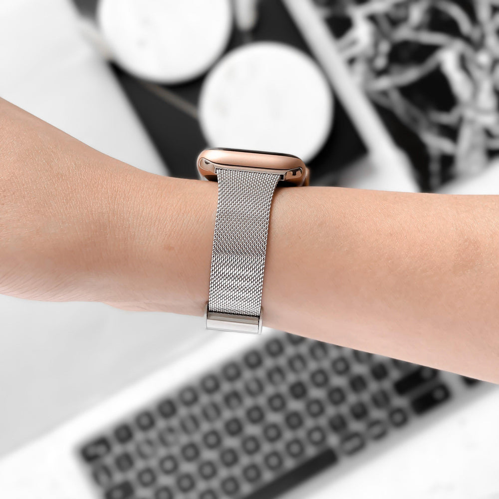 A close-up of a person's wrist wearing a smartwatch with a Coconut Lane Milanese Apple Watch Strap in Silver. In the blurred background, there is a keyboard and some round objects on a white surface with black patterns.