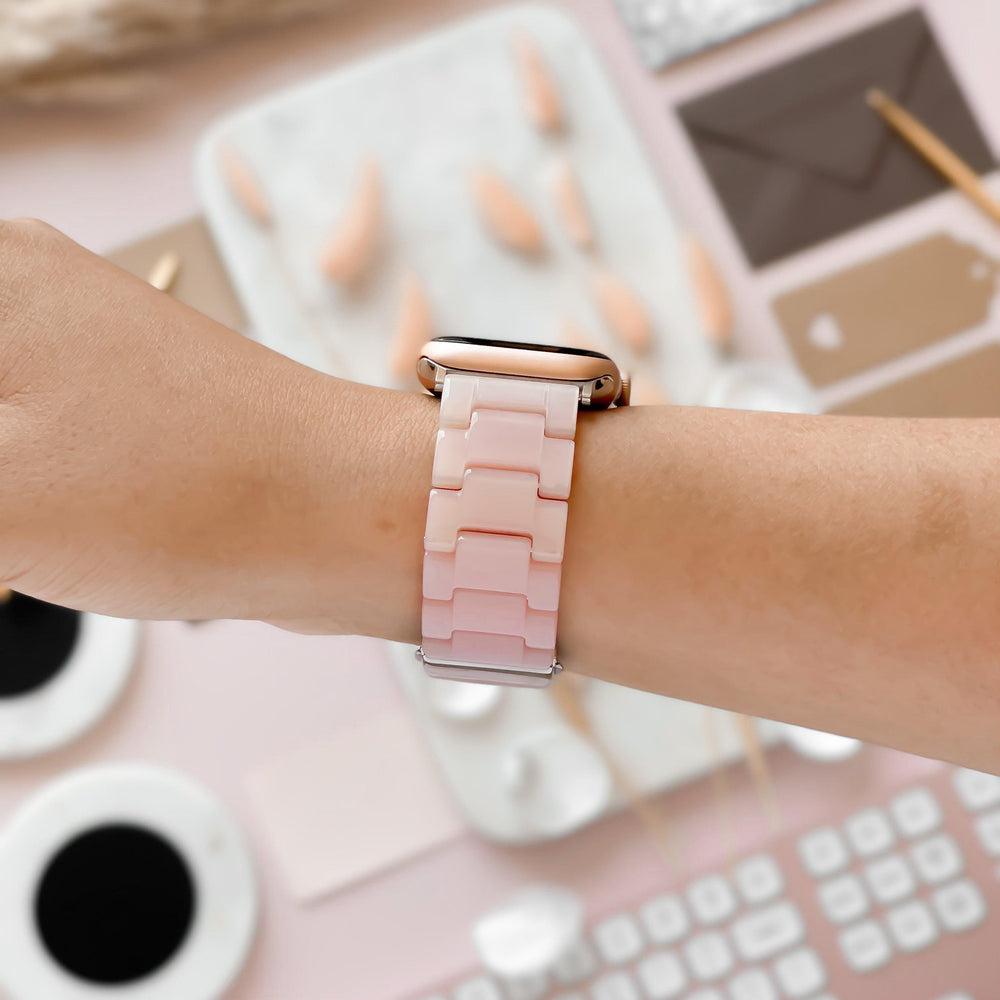 A close-up of a wrist wearing a smartwatch with a Luxe Pink Apple Watch Strap from Coconut Lane. The background includes various objects such as a keyboard, coffee cups, stationery, and decorative items, all arranged on a pastel-colored workspace.