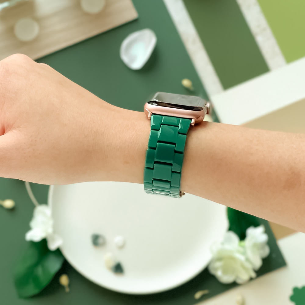 A person wears a Coconut Lane Luxe Green Apple Watch Strap with a glossy finish on their wrist. In the background, a green table displays various items, including a white plate, flowers, and decorative stones. The scene is bright and well-lit.