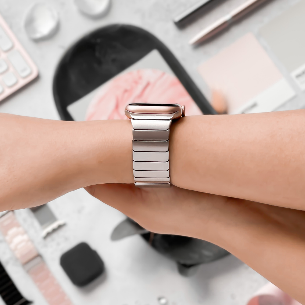 Close-up of a hand wearing a sleek, modern smartwatch with an adjustable Slinky Apple Watch Strap - Silver by Coconut Lane. The background is in soft focus and includes various items such as pastel-colored swatches, a keyboard, and other small accessories. The FREE link remover tool allows for easy customization.
